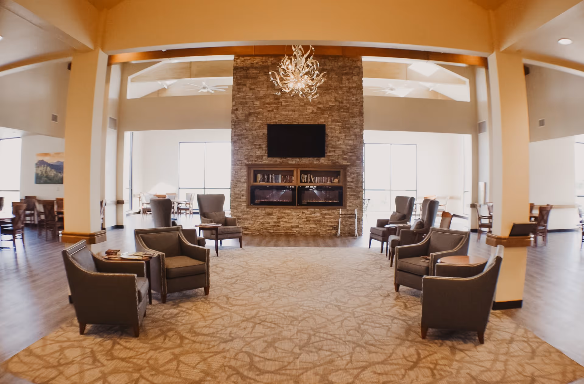 Spacious and well-lit common area with a large stone fireplace and a mounted flat-screen TV above it. The room features several comfortable armchairs arranged in pairs around small tables on a patterned carpet. Large windows on either side of the fireplace allow natural light to fill the space. In the background, there are dining tables and chairs on a wooden floor, with high ceilings and ceiling fans visible.