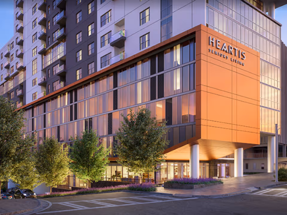 Exterior view of the Heartis Buckhead Senior Living building at dusk, featuring a modern multi-story structure with large glass windows, an orange accent panel with the facility name, and landscaped greenery in front.