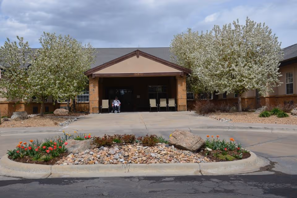 Front entrance of a senior living building with a circular rock-and-flower island, blossoming trees, and a covered drive-up entry.