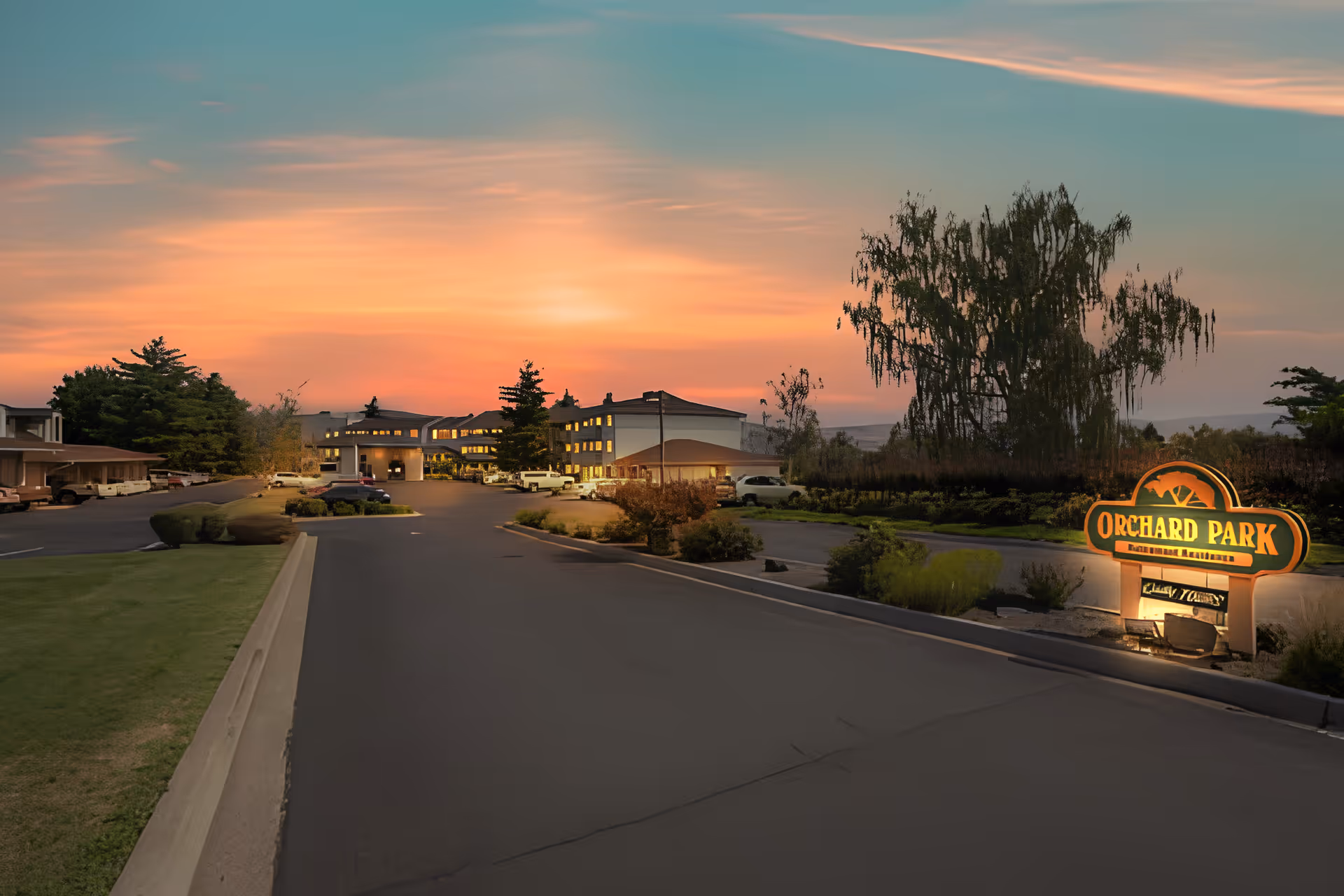 Driveway leading to a lit multi-story senior living building at sunset with an illuminated "Orchard Park" entrance sign.