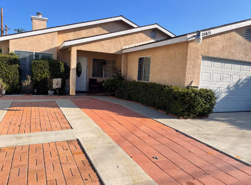 Single-story beige senior housing building with brick driveway, hedges, and front entrance.