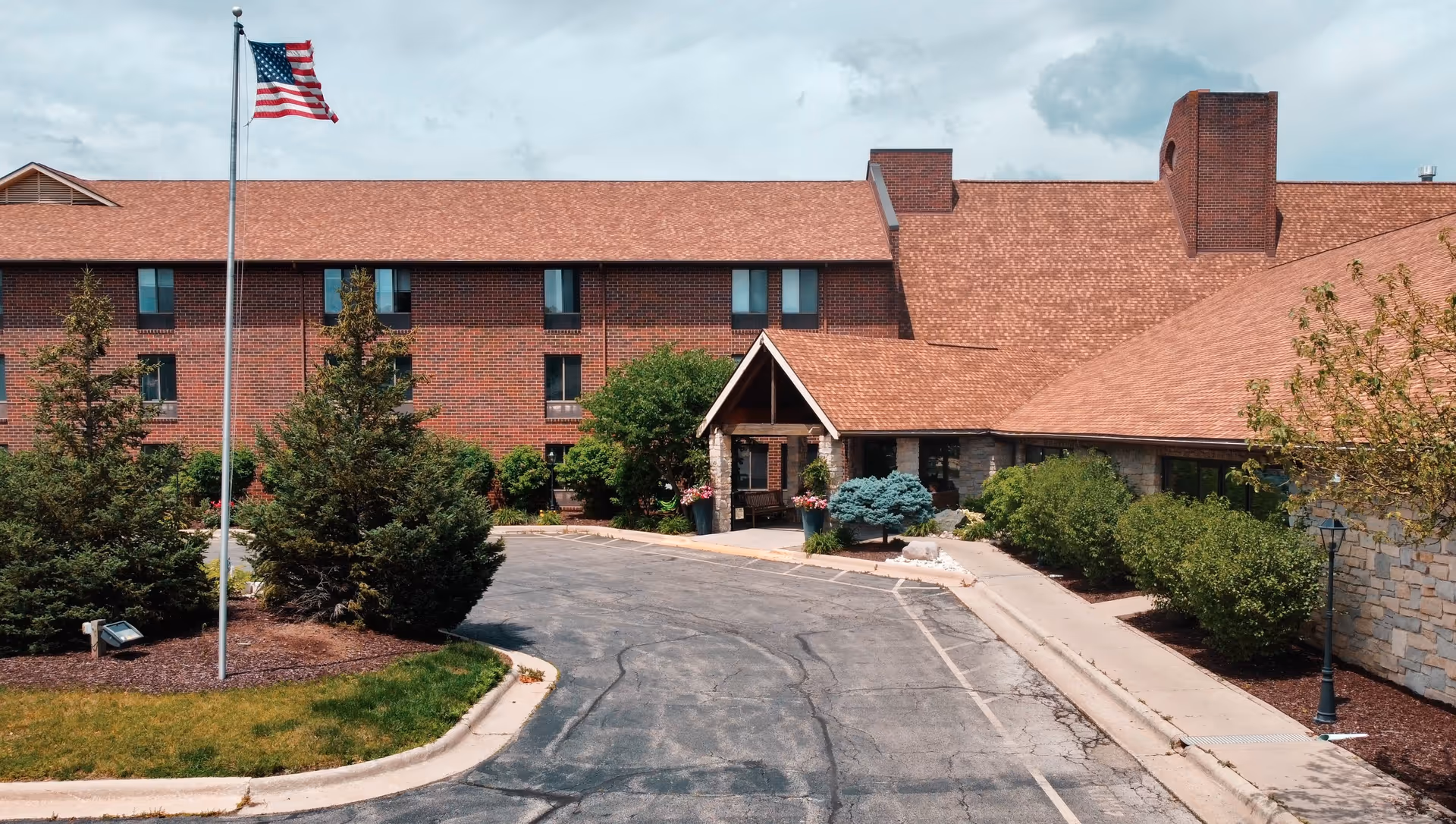 Exterior view of a senior living facility named Independence Village with a brick building, a driveway, landscaped bushes and trees, and an American flag on a flagpole.