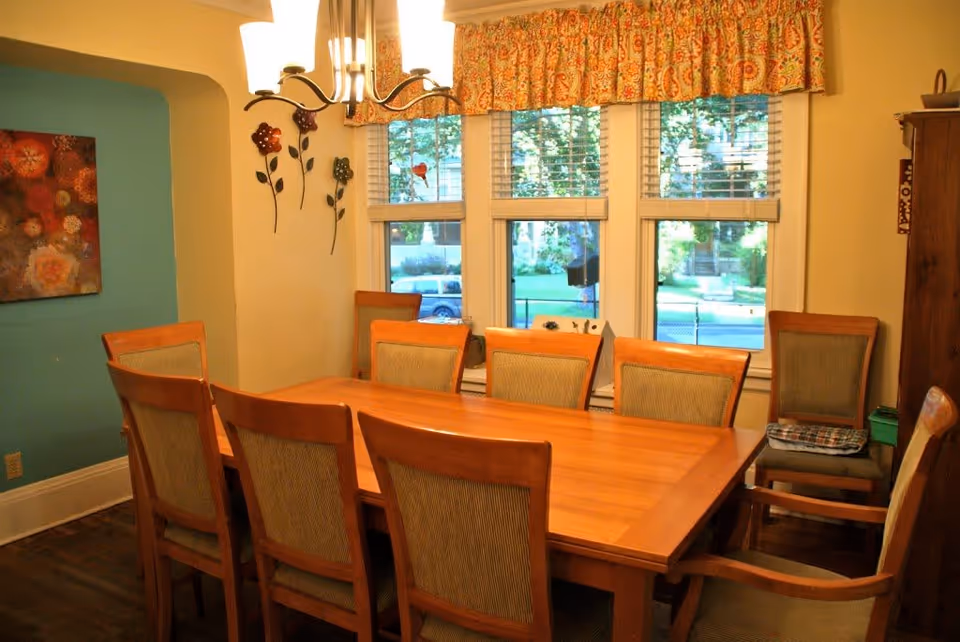 A dining room with a large wooden table surrounded by eight cushioned chairs. There is a chandelier hanging above the table, a window with floral valance curtains letting in natural light, and wall decorations including a floral painting and metal flower art.