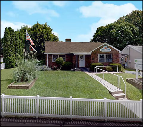 Front view of a single-story brick senior care facility with a white fence and green lawn.