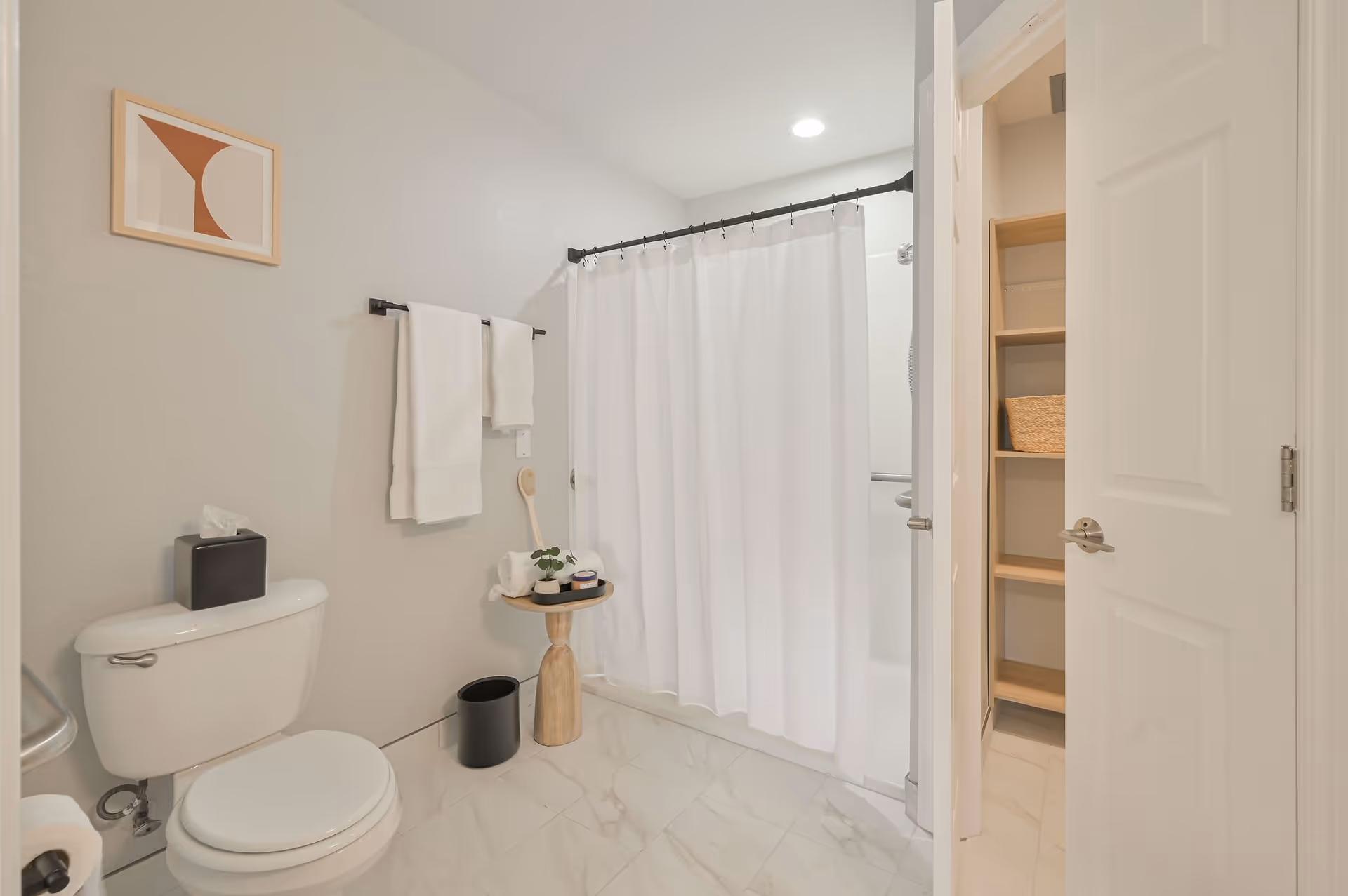 A clean, modern bathroom with a white toilet, a black tissue box on the toilet tank, white towels hanging on a black towel rack, a small wooden side table holding rolled towels and toiletries, a white shower curtain covering a bathtub, and an open door revealing wooden shelves with a wicker basket.