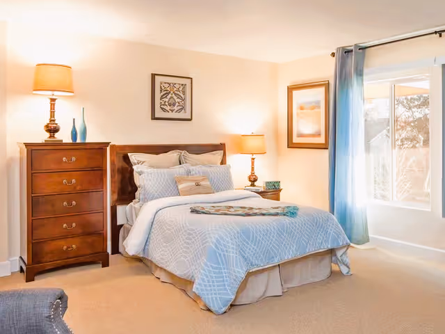 A neatly made bedroom with a bed, dresser, lamps, and large window.