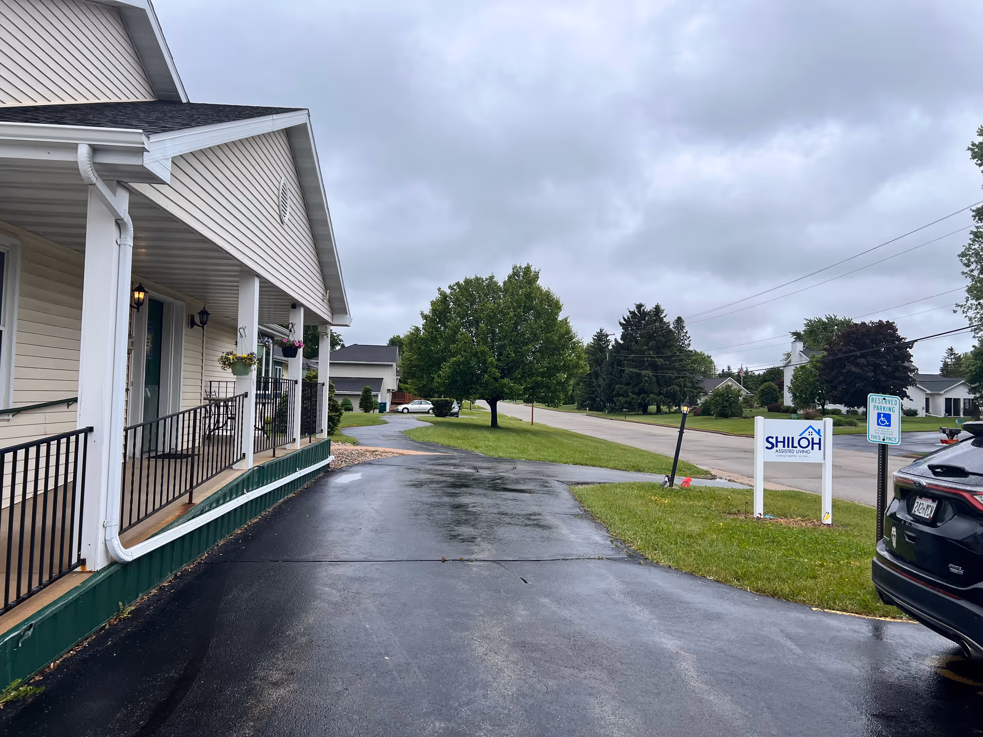 Front entrance and covered porch of Shiloh Assisted Living with a driveway, facility sign, parked car, and overcast sky.