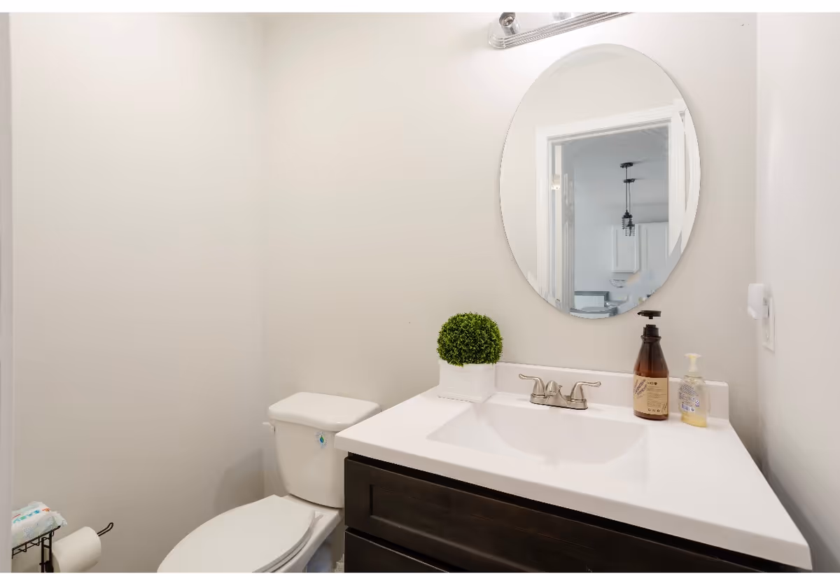 A small bathroom with a white toilet and a white sink countertop with a dark brown cabinet. On the countertop, there is a small green potted plant, a brown soap dispenser, and a clear hand soap dispenser. Above the sink is a round mirror reflecting part of an adjacent room with white cabinets and a hanging light fixture.