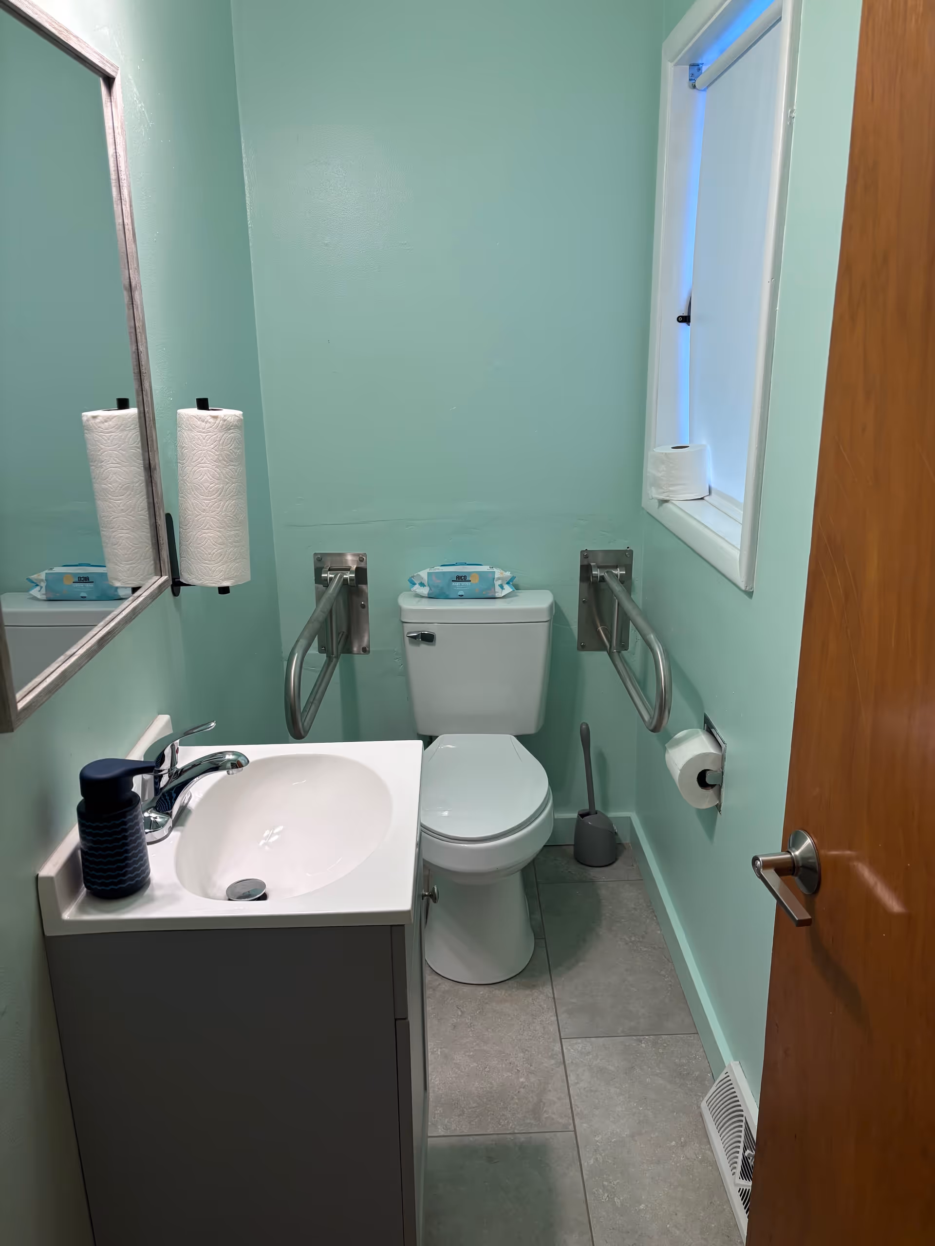 Small mint-green bathroom with sink, toilet, grab bars, toilet paper, and a window.