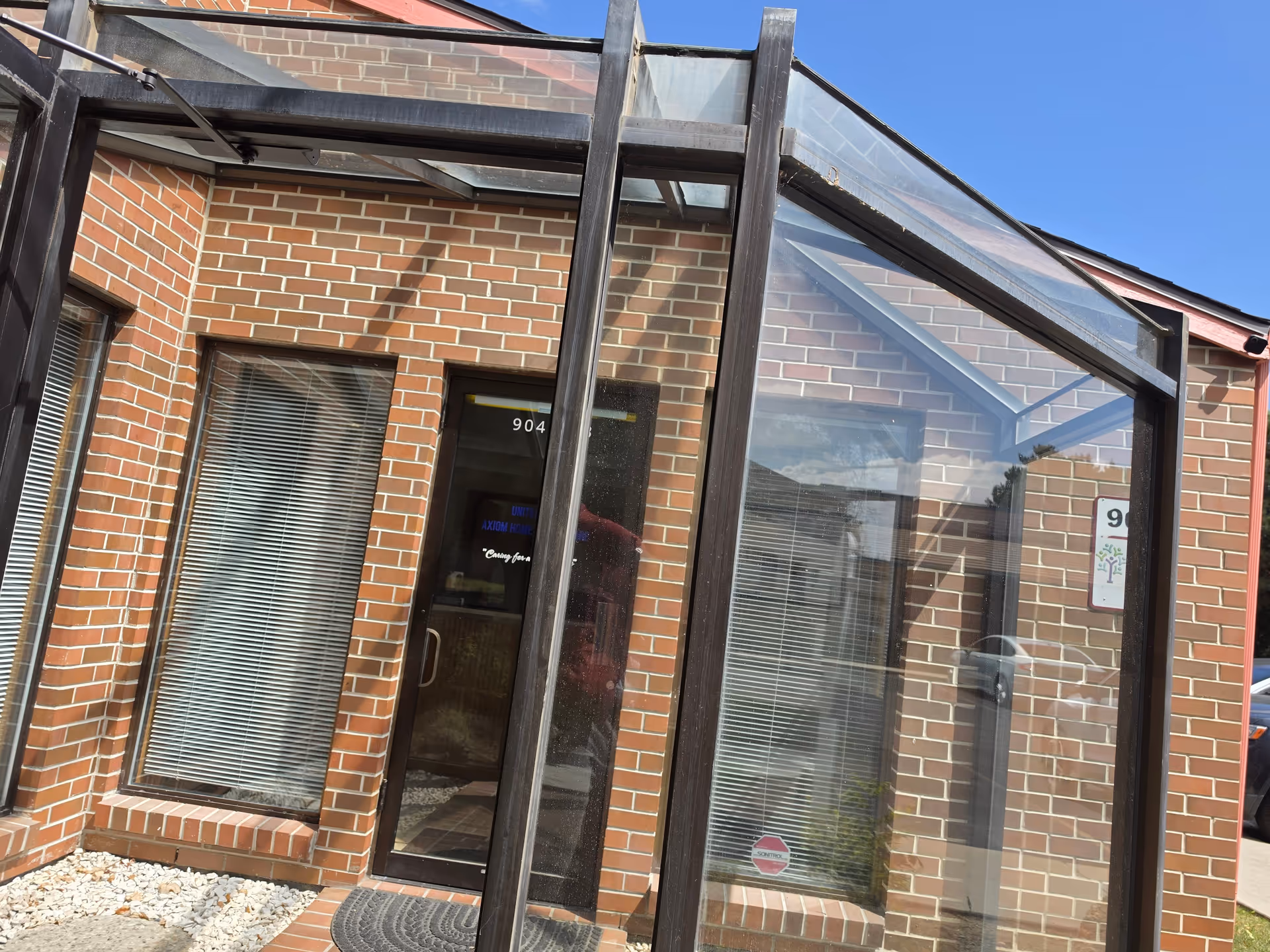 Brick facility entrance with glass-enclosed foyer and metal framework under blue sky.