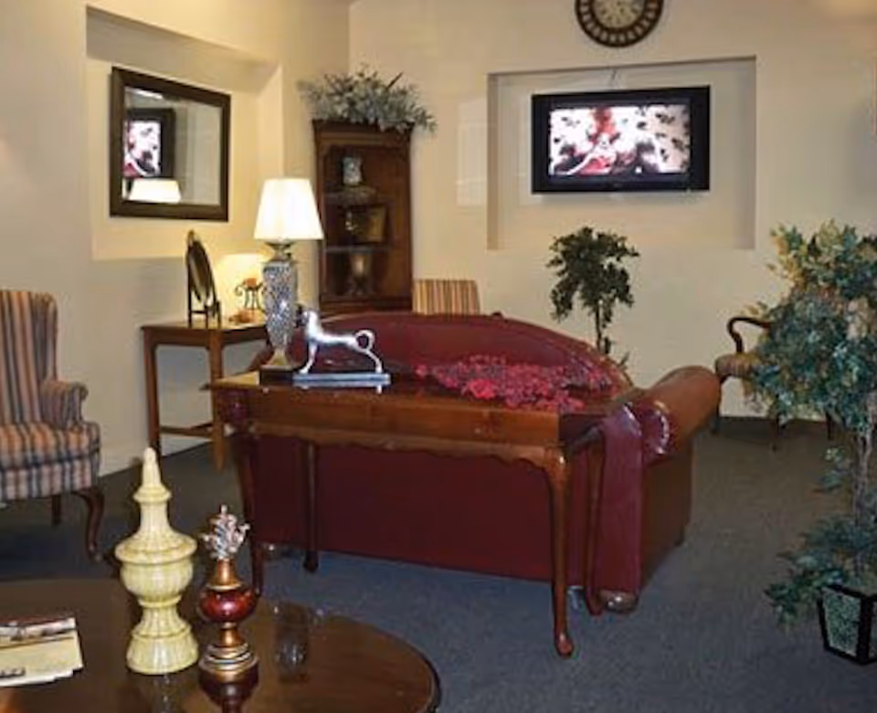 Cozy common room with a red upholstered sofa, armchairs, a side table with a lamp and decorative items, and a wall-mounted TV.