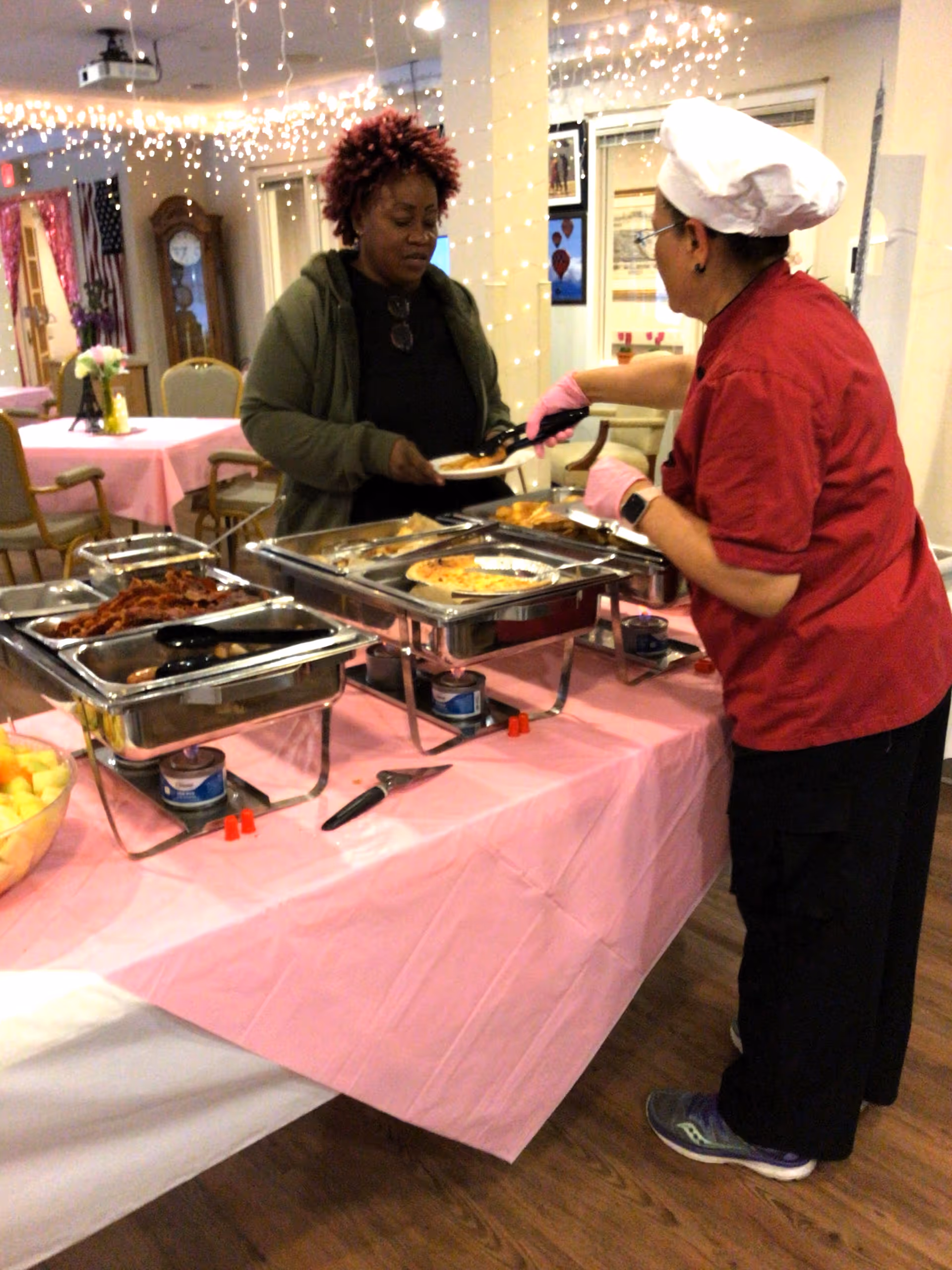 A server in red uniform serves food at a pink-draped buffet in a senior dining room.