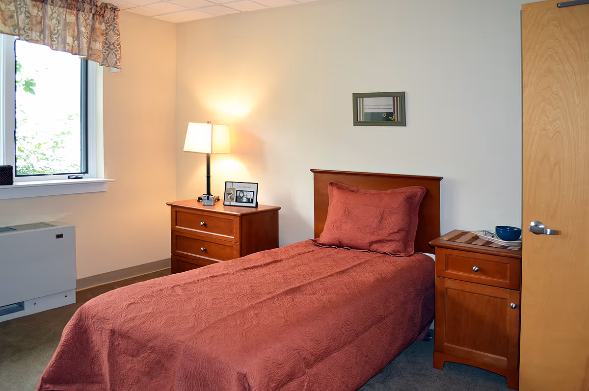 A small bedroom with a single bed covered in a reddish-brown quilt and matching pillow. There are two wooden nightstands on either side of the bed, one with a lamp and a framed photo, the other with a bowl and placemat. A window with a floral valance lets in natural light, and a wooden door is partially visible on the right side of the image.