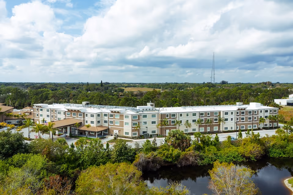 Aerial view of a three-story senior living building (Discovery Village at Stuart) surrounded by trees and a pond under a cloudy sky.