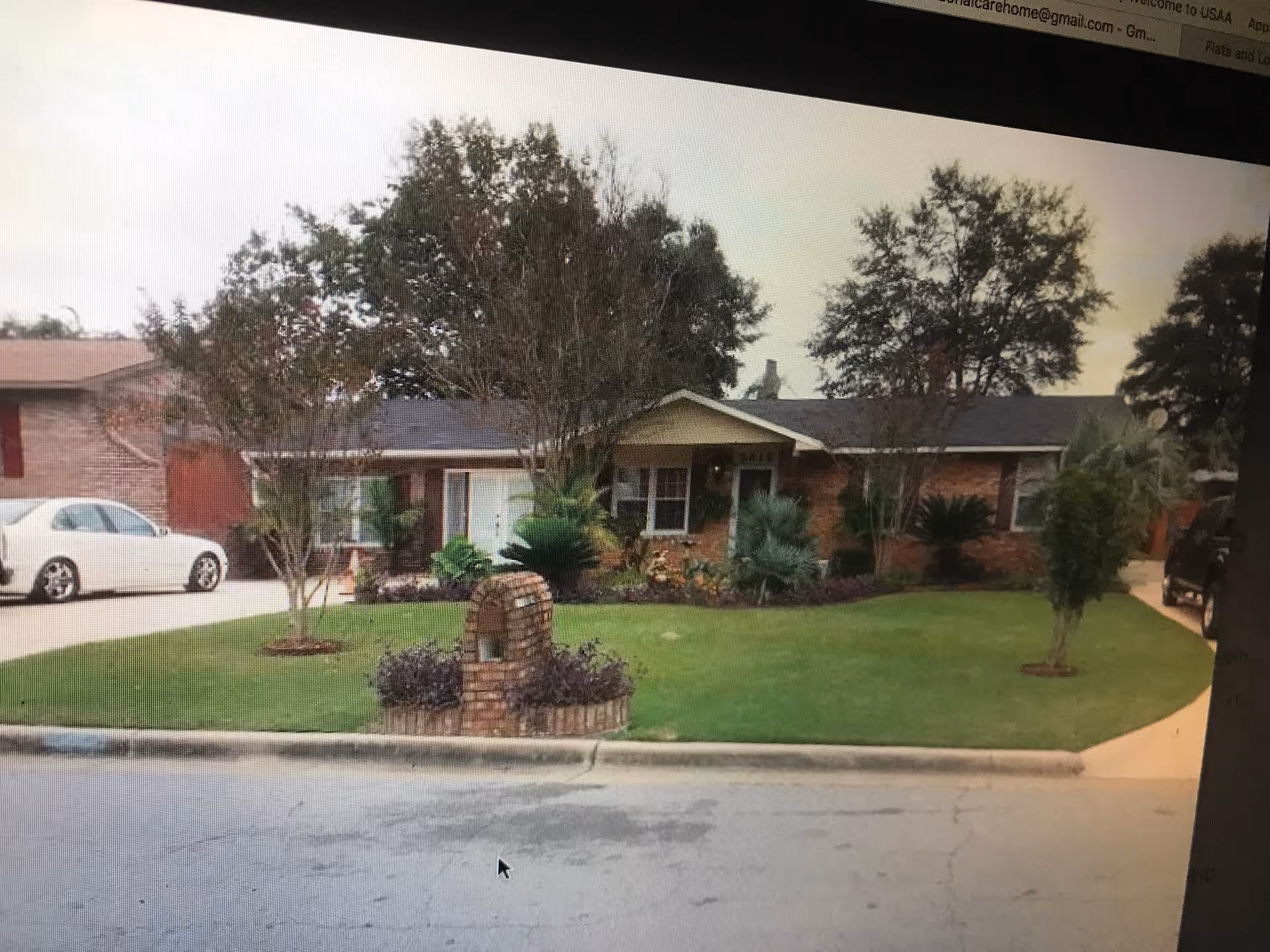 Front exterior view of a single-story brick house with a well-maintained lawn, several trees, shrubs, and a brick mailbox. There are two cars parked on either side of the house driveway.