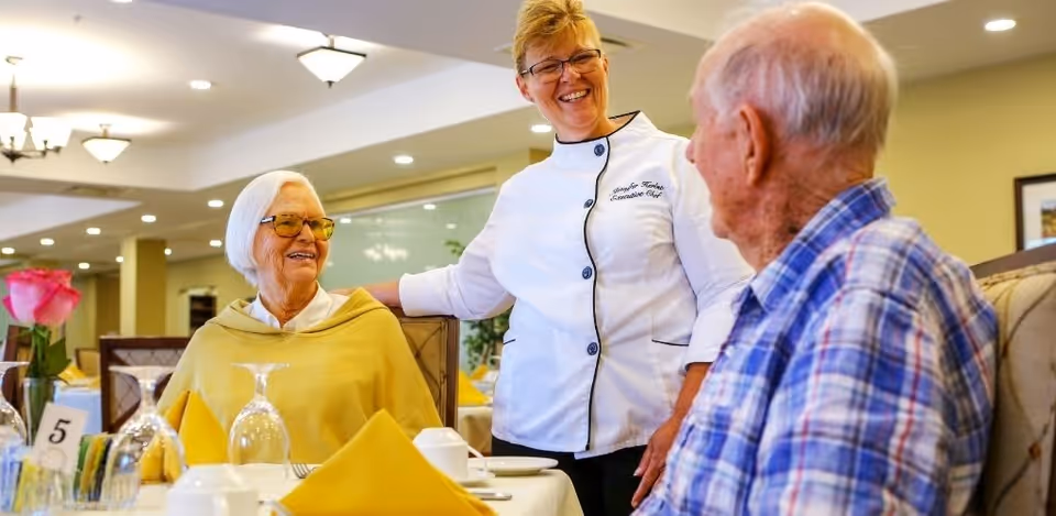 A smiling chef in a white uniform stands and talks with two elderly people seated at a dining table set with yellow napkins, glassware, and a pink rose in a vase in a well-lit dining room.