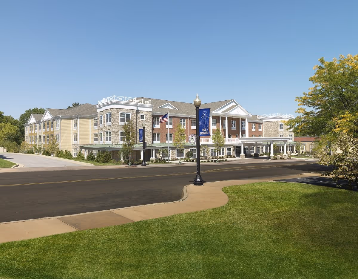 Front exterior of a multi-story senior living facility with columns, flags, and landscaped lawn under a clear blue sky.
