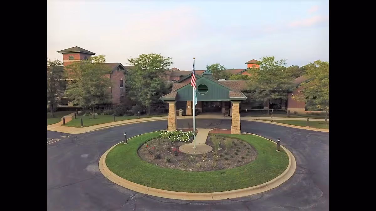Front exterior view of Valley Hi Nursing & Rehabilitation for McHenry County showing a circular driveway with a landscaped island in the center featuring a flagpole with the American flag and another flag. The building is a multi-story brick structure with green roofing and surrounded by trees.