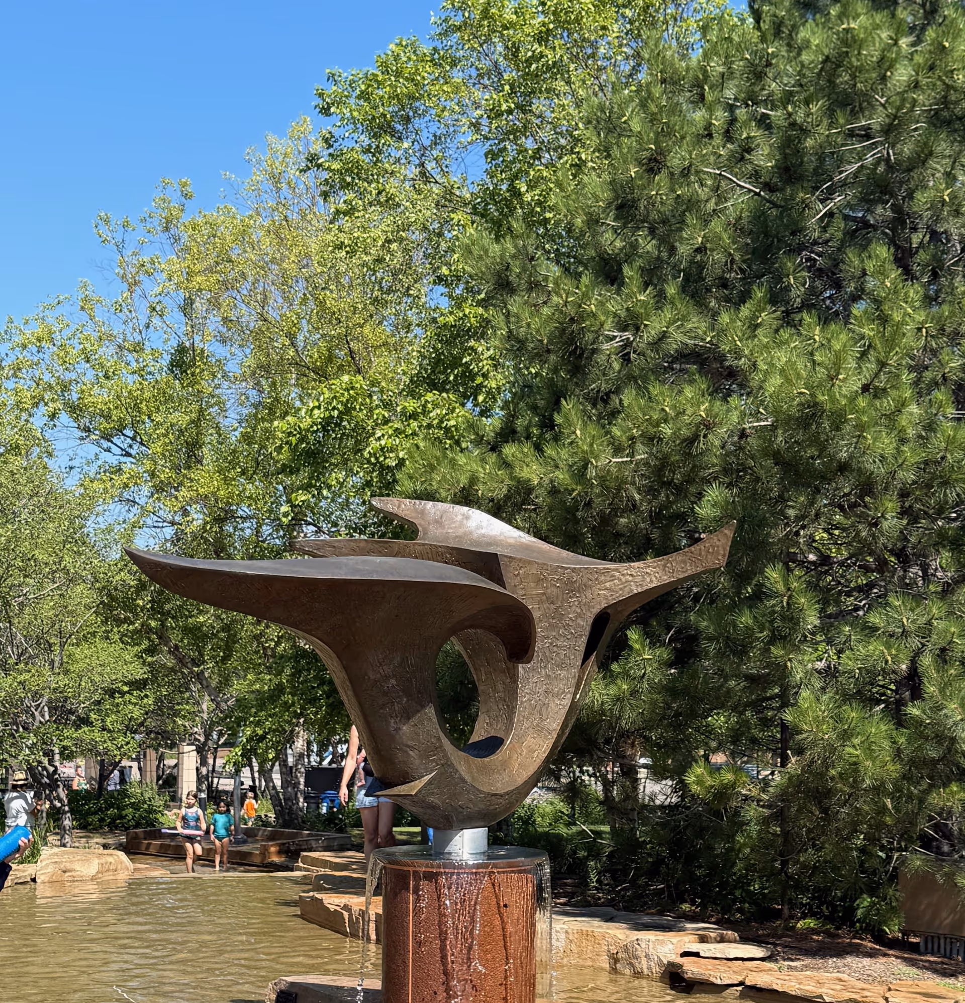 Abstract bronze sculpture in a sunny outdoor courtyard with trees and a water feature.