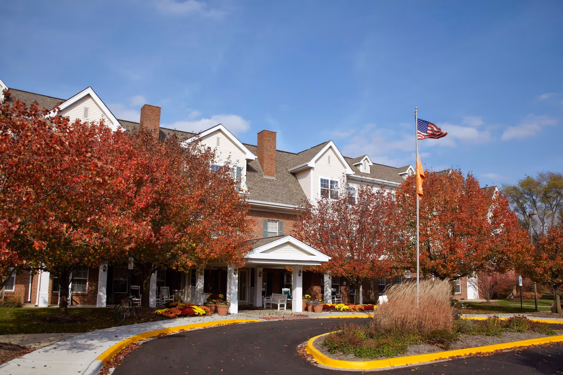 Front exterior of a multi-story senior living building with autumn trees, an American flag, and a circular driveway.