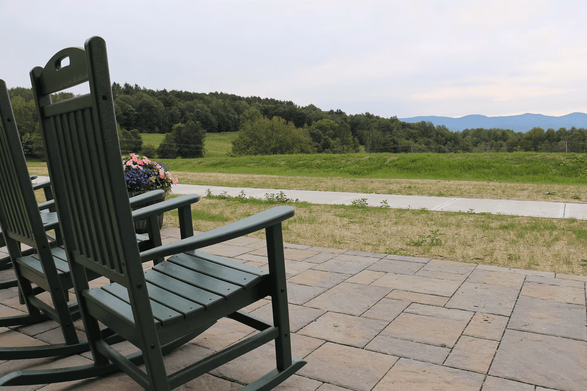 Rocking chairs on a stone patio overlook a grassy field with distant hills.