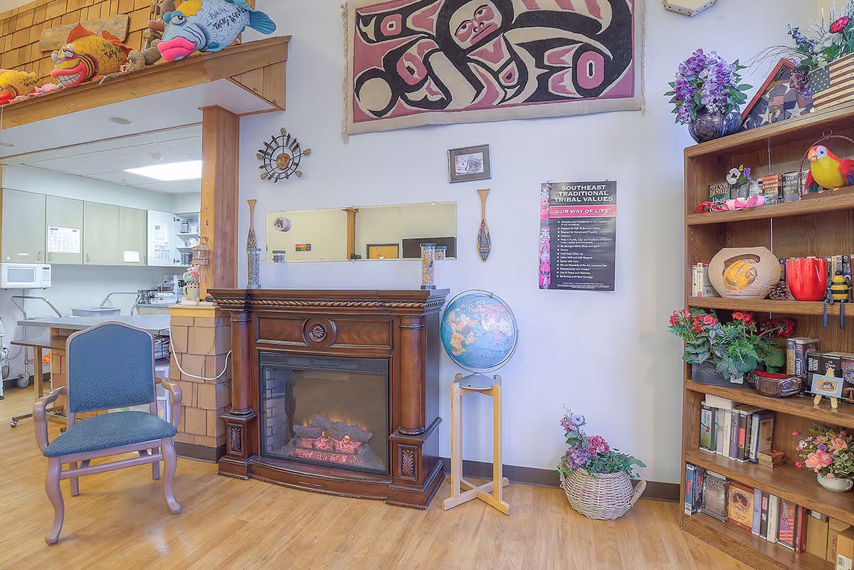 A cozy communal room showing a wooden electric fireplace, upholstered chair, globe on a stand, and a bookshelf beside a kitchen pass-through.