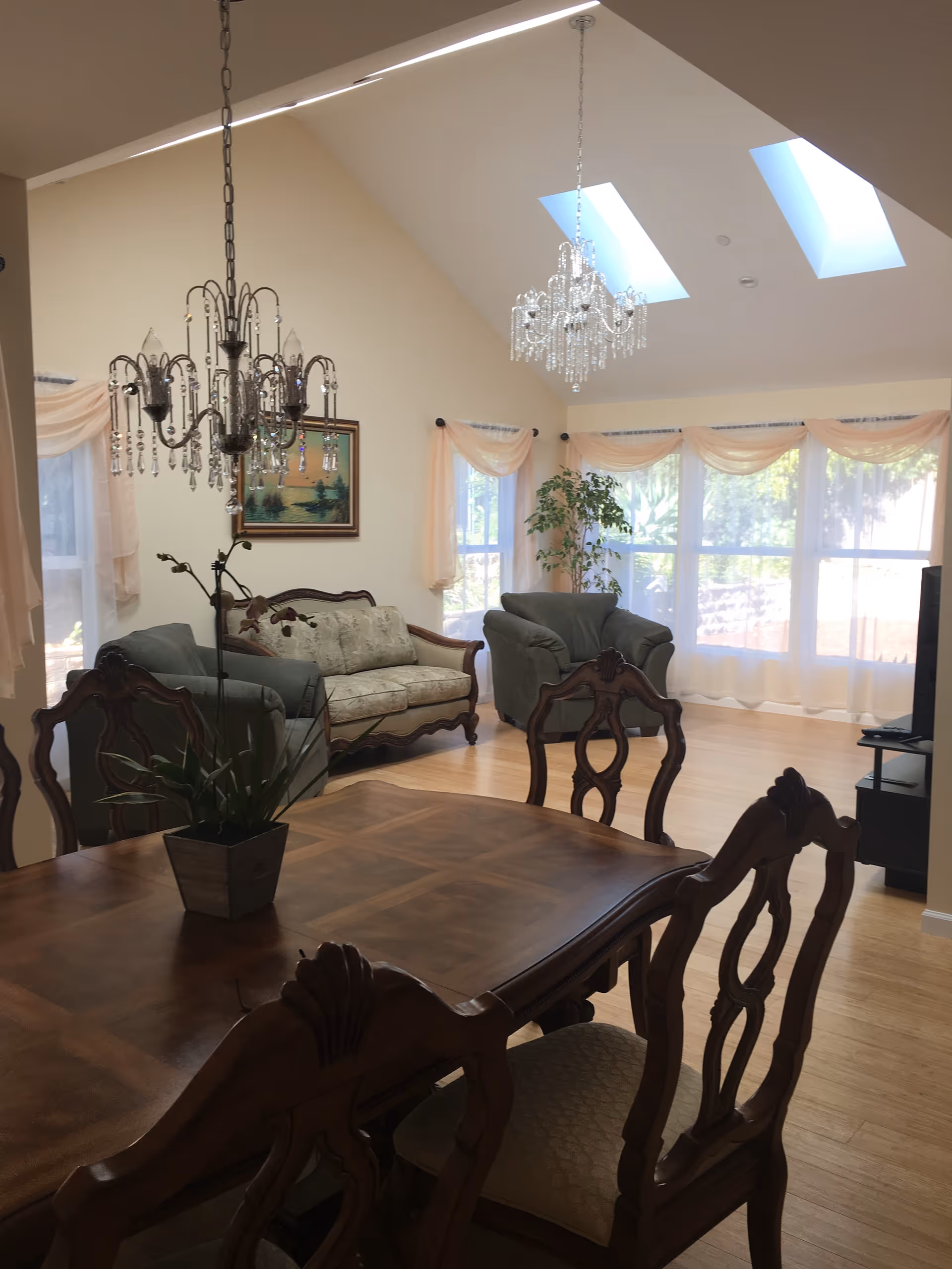 A bright living and dining area with wooden dining table and chairs in the foreground, two chandeliers hanging from a vaulted ceiling with skylights, a sofa and armchair near large windows with sheer curtains, and a potted plant in the corner.