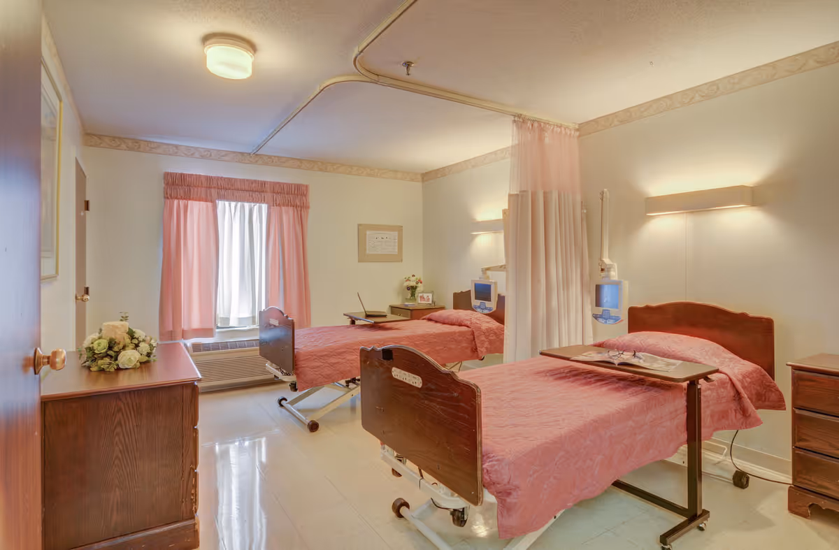 Two pink hospital beds in a curtained, well-lit bedroom with bedside tables and window drapes.
