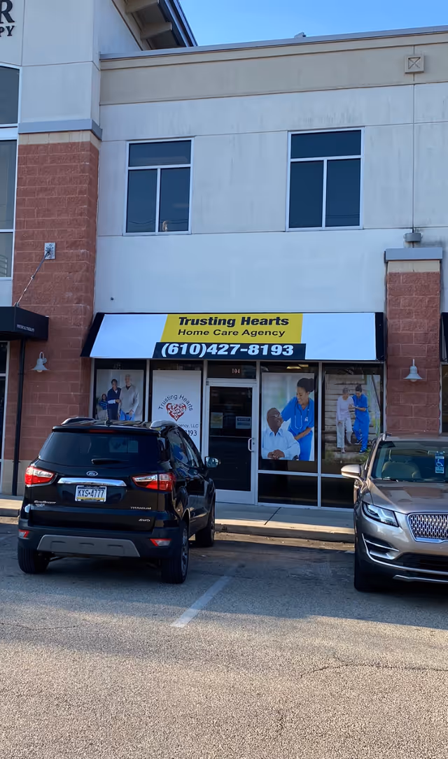 Front of Trusting Hearts Home Care Agency storefront with a yellow sign and parked cars.