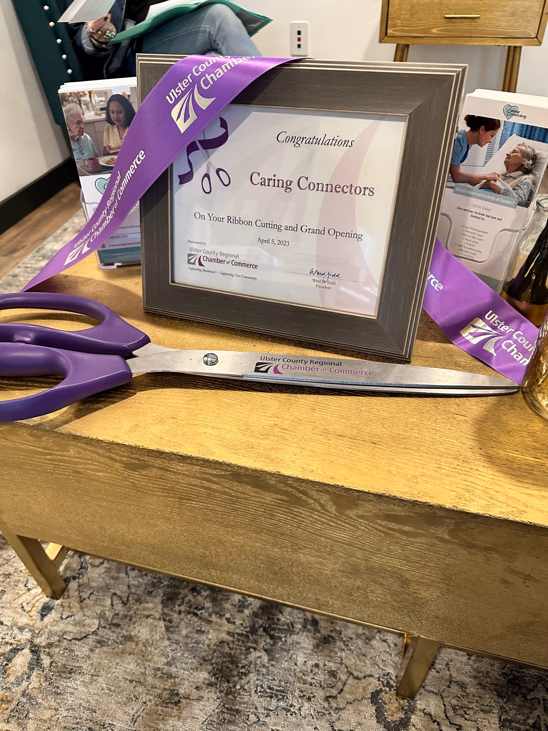Framed Caring Connectors ribbon-cutting certificate on a desk with purple scissors