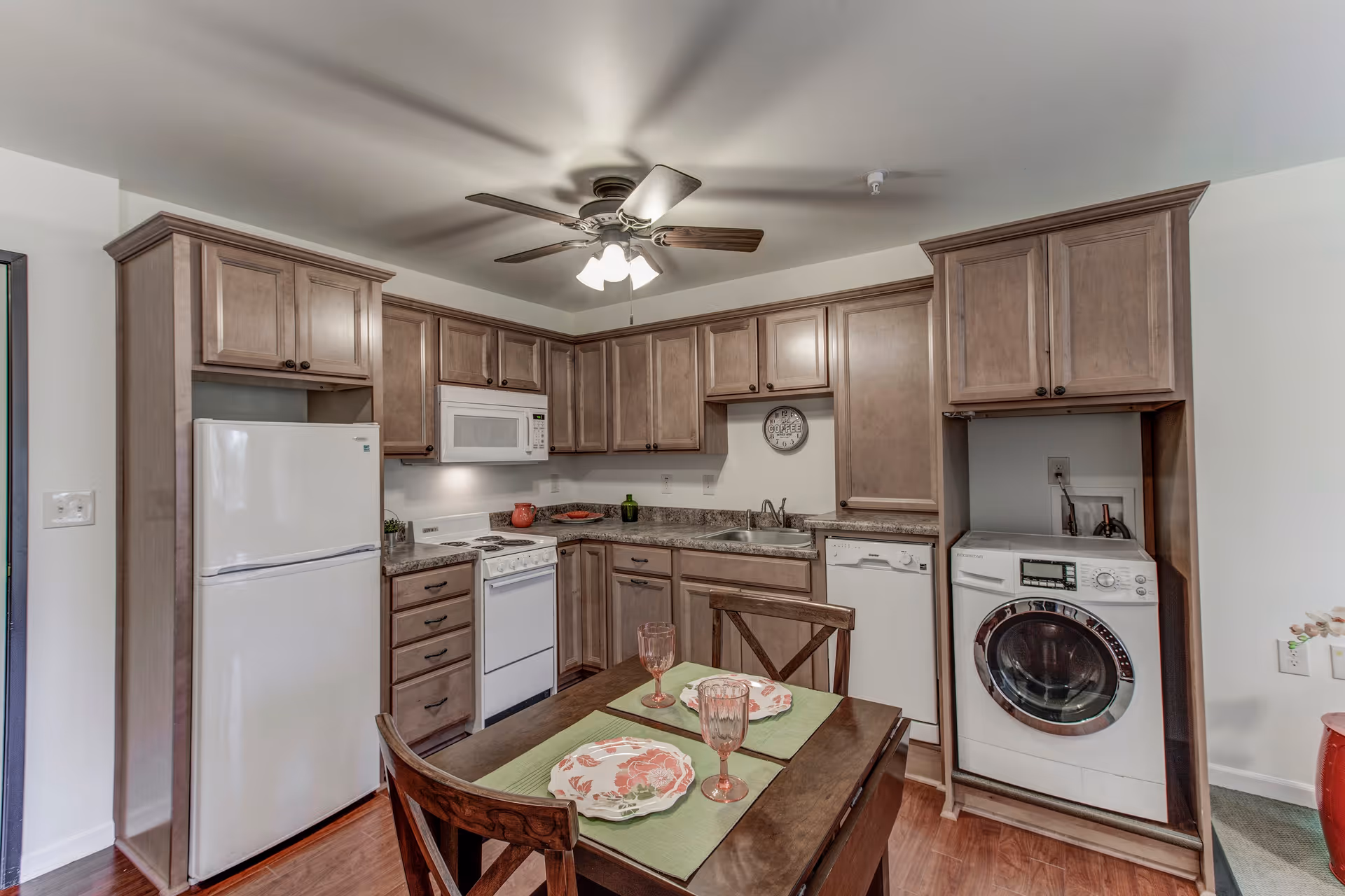 A compact kitchen with wooden cabinets, a white refrigerator, microwave, stove, dishwasher, and a washing machine. A small dining table with two chairs is set with plates and glasses. A ceiling fan with lights is mounted above.