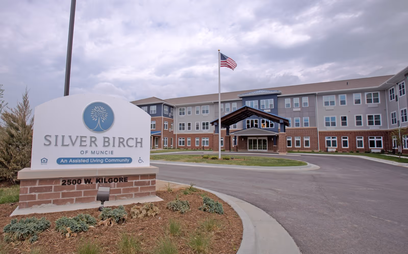 Exterior view of Silver Birch of Muncie, an assisted living community building with a large sign in the foreground displaying the facility name and address 2500 W. Kilgore. The building is three stories tall with multiple windows, a covered entrance, and an American flag on a flagpole in front. The sky is cloudy.