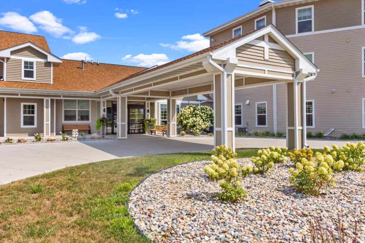 Exterior view of a senior living facility entrance with a covered drop-off area, beige siding, white trim, and a landscaped garden with flowering bushes and rocks in front.