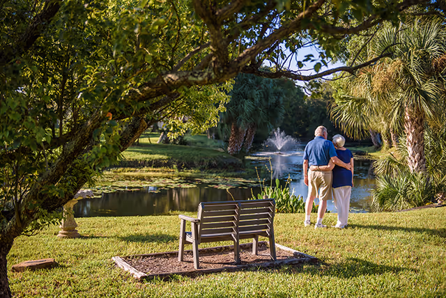 An elderly couple arm-in-arm stands by a pond with a fountain in a landscaped garden with a bench and trees.