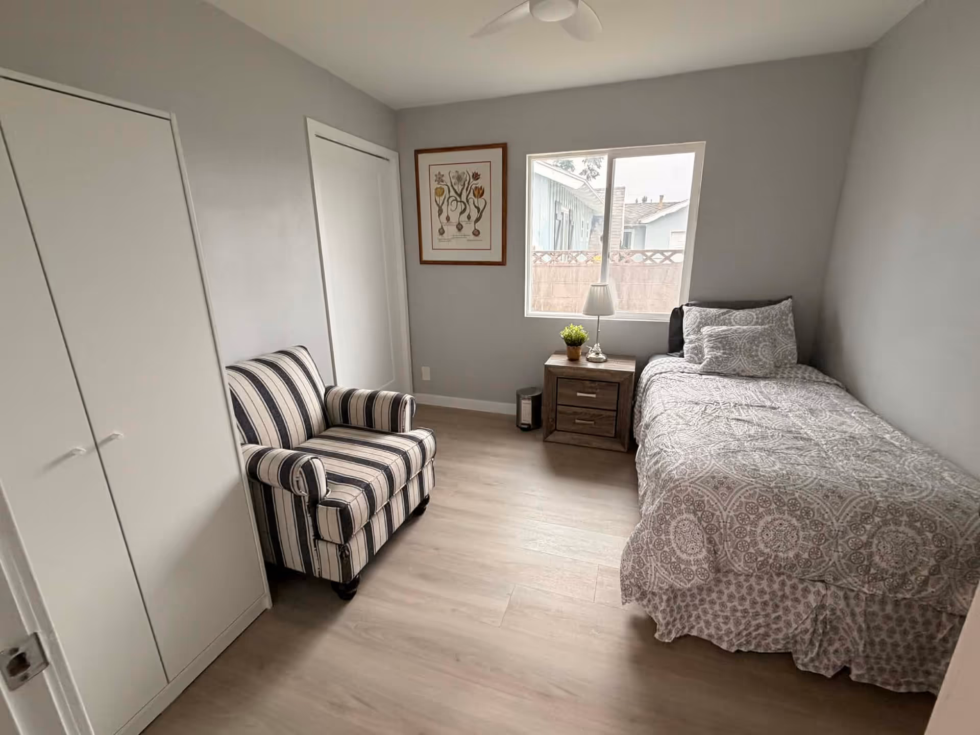 A small bedroom with a bed, nightstand, striped armchair, and closet, bright window.