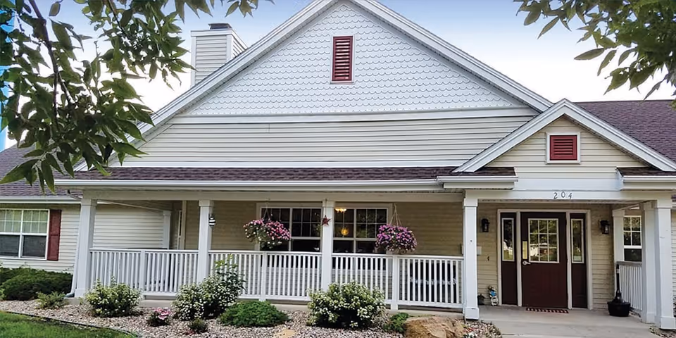 Front exterior view of a single-story senior living facility building with beige siding, white trim, and maroon shutters. The building has a covered porch with white railings, hanging flower baskets, and a maroon front door with sidelights. The landscaping includes small bushes and flowers in a rock bed.