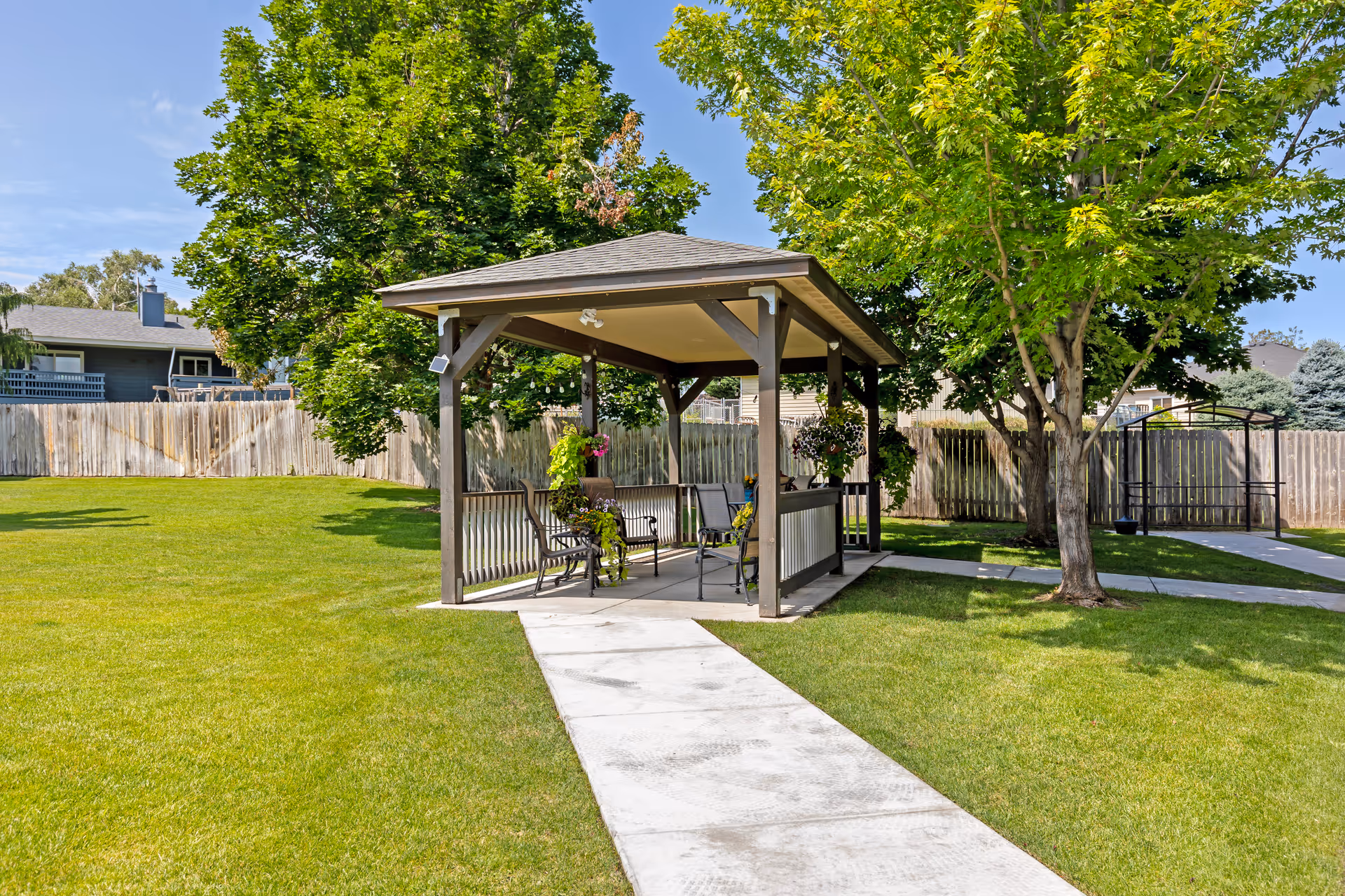 A small outdoor gazebo with a roof and open sides, containing benches and chairs, surrounded by green grass, trees, and a wooden fence in the background under a clear blue sky.