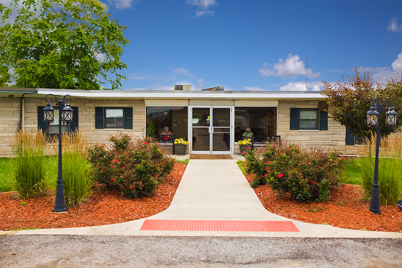 Front exterior view of a single-story building with a concrete walkway leading to a glass entrance door. The building has beige stone walls, green shutters on the windows, and is surrounded by landscaped bushes, grass, and lamp posts under a blue sky with some clouds.