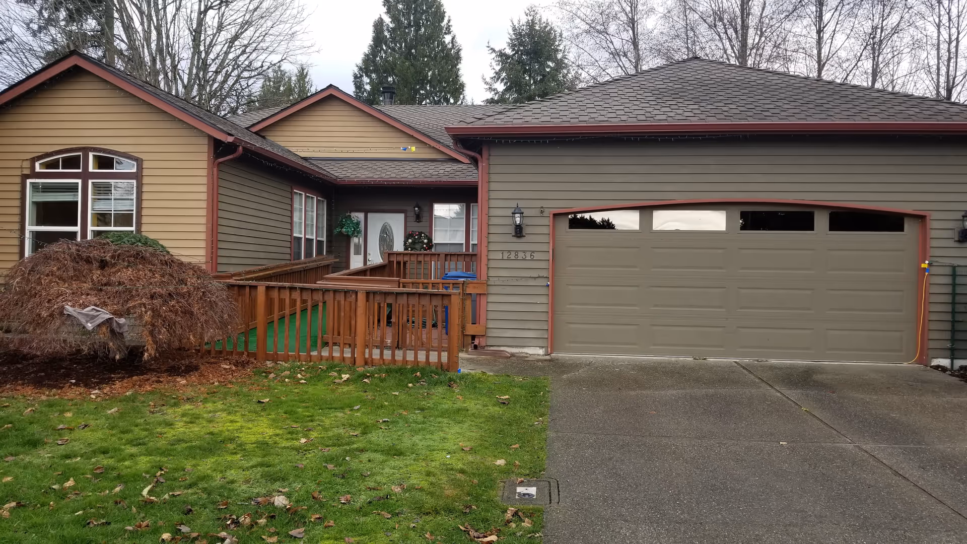 Exterior view of a single-story residential building with a two-car garage, a wooden ramp leading to the front door, and a small lawn with a tree in front. The house has brown siding with red trim and several windows.