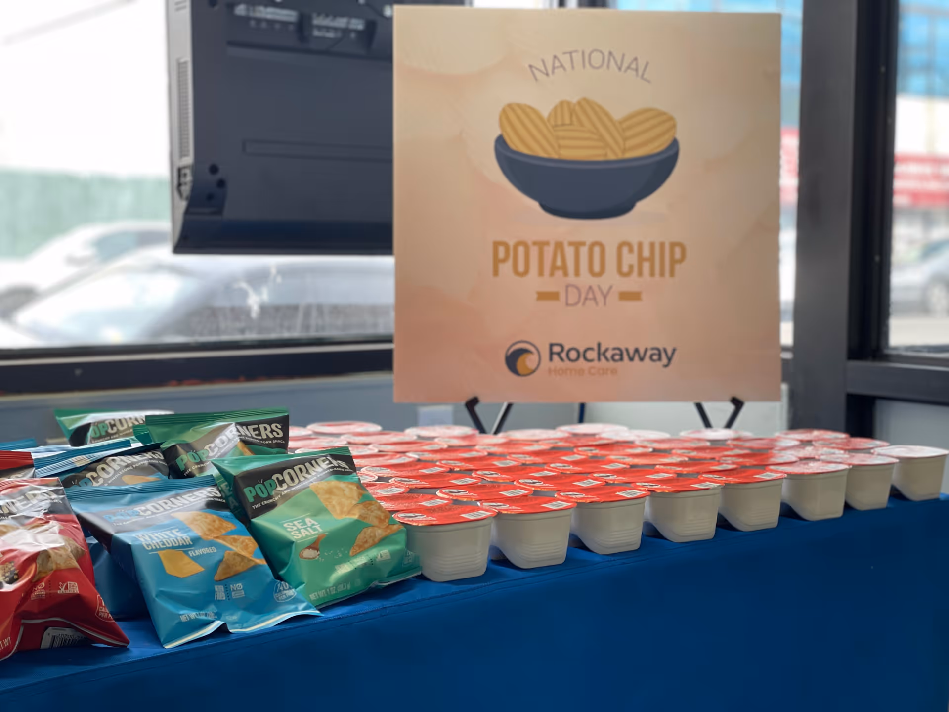 Indoor display table with snack bags and cups beside a National Potato Chip Day sign.