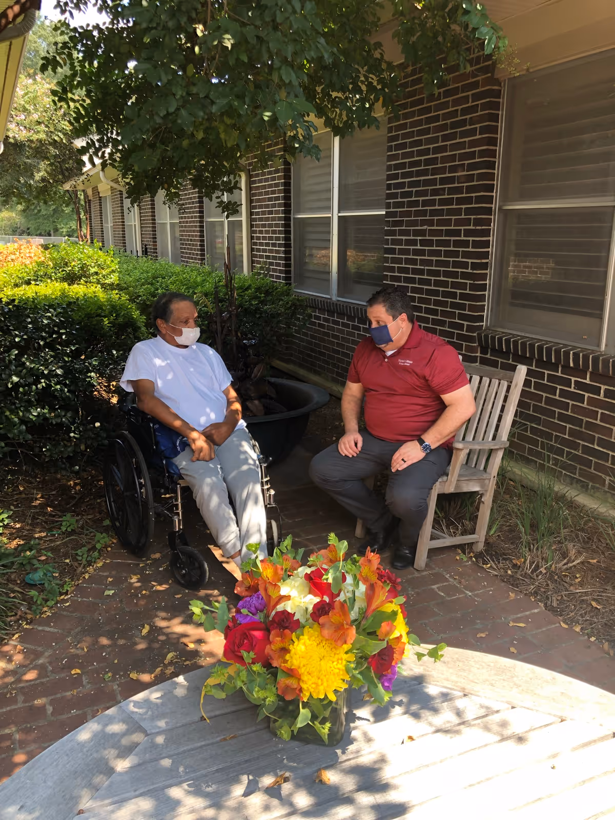 Two people wearing face masks are sitting outside near a brick building. One person is in a wheelchair wearing a white shirt and light-colored pants, while the other person is seated on a wooden chair wearing a red polo shirt and dark pants. In the foreground, there is a wooden table with a colorful bouquet of flowers. Green bushes and a tree provide shade in the background.