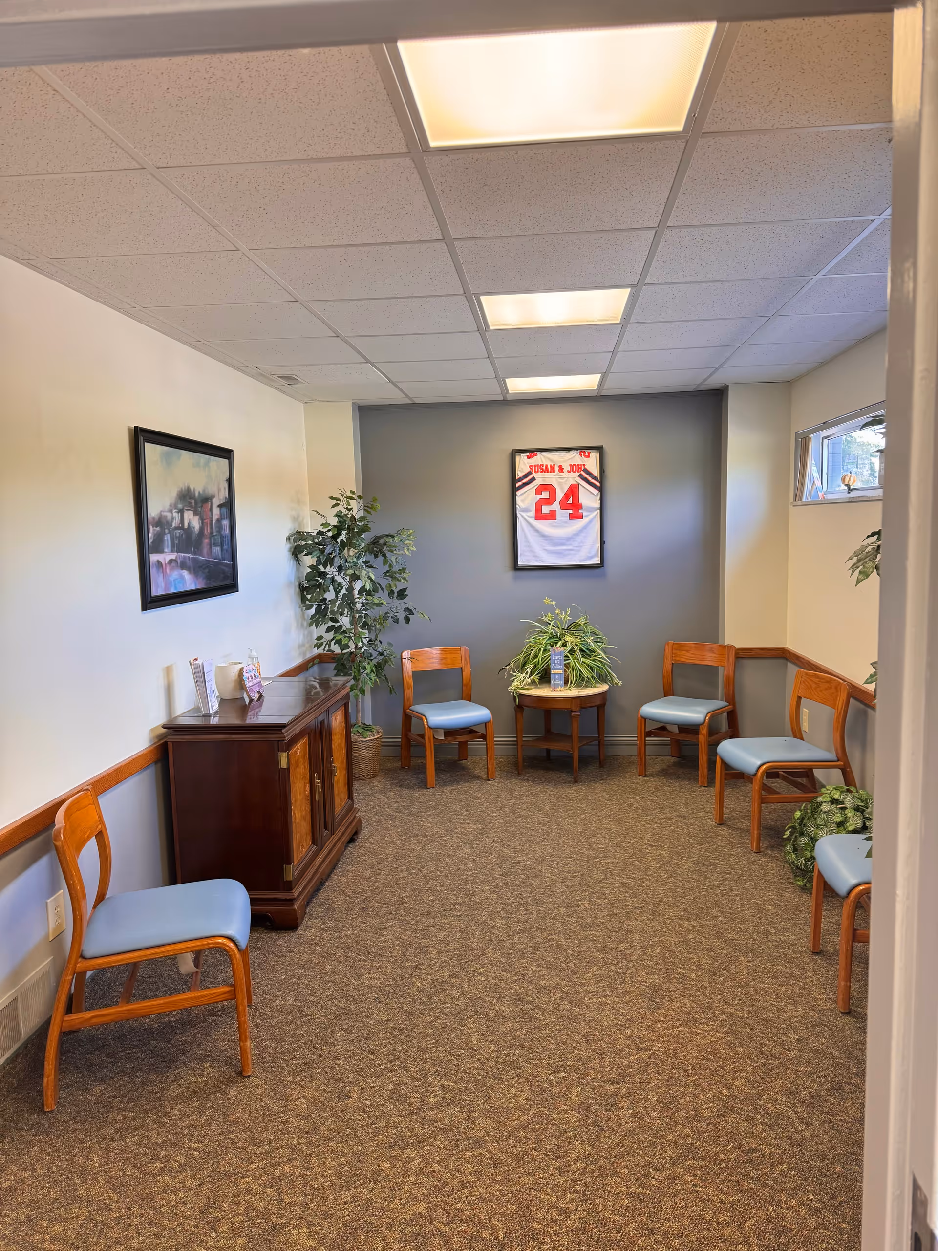 Small lounge room with blue-cushioned chairs, plants, and a framed jersey on the wall.