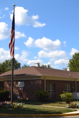 Single-story brick senior living building with an American flag on a pole and landscaping under a blue sky.