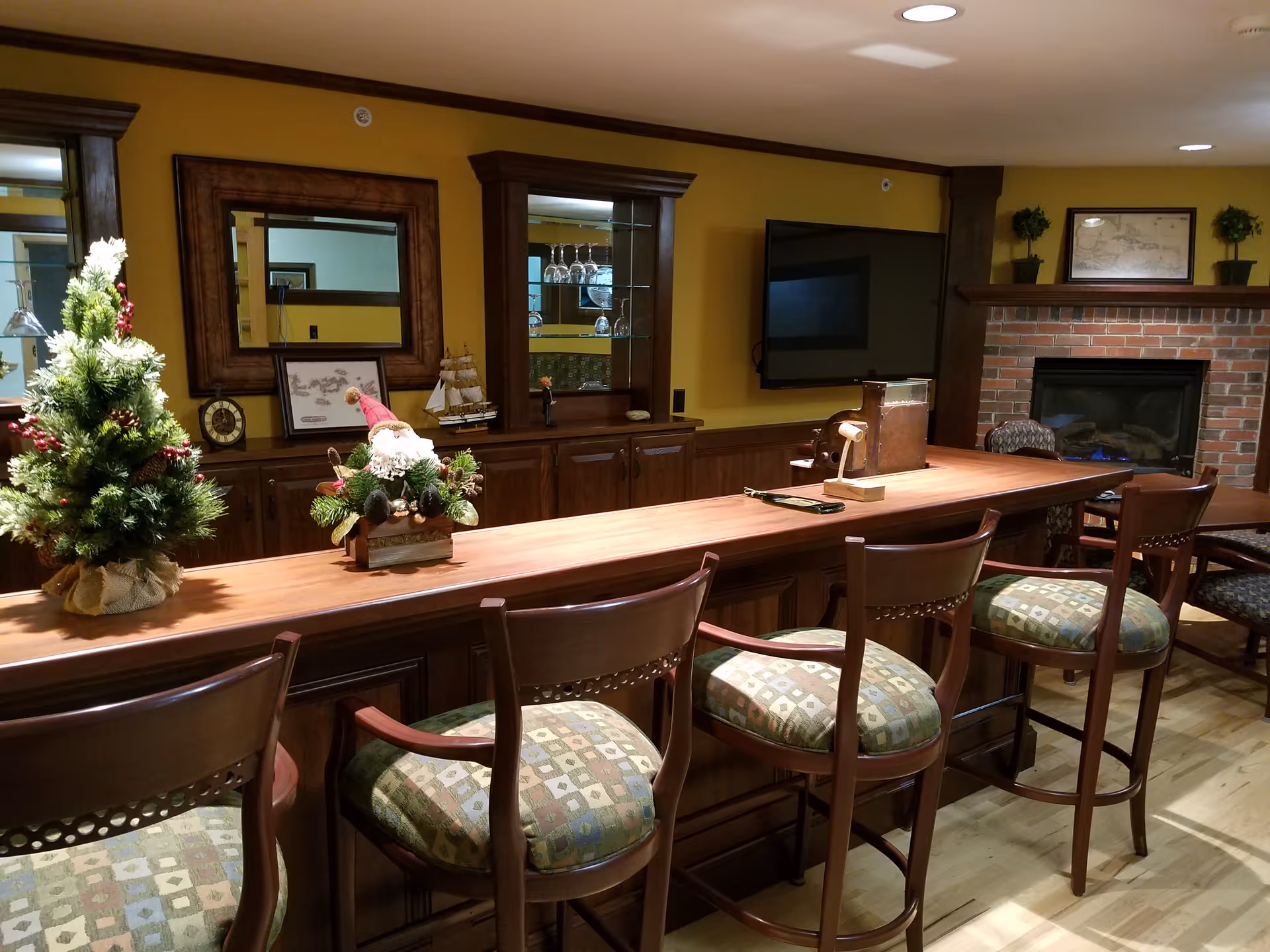 A cozy interior room with a long wooden bar counter and four cushioned bar stools. On the counter, there are small decorative items including a miniature Christmas tree and a Santa Claus figure. Behind the bar, there is a wooden cabinet with glass shelves holding glassware, a wall-mounted TV, and a brick fireplace with a mantel decorated with framed artwork and small potted plants. The walls are painted yellow and the floor is wooden.