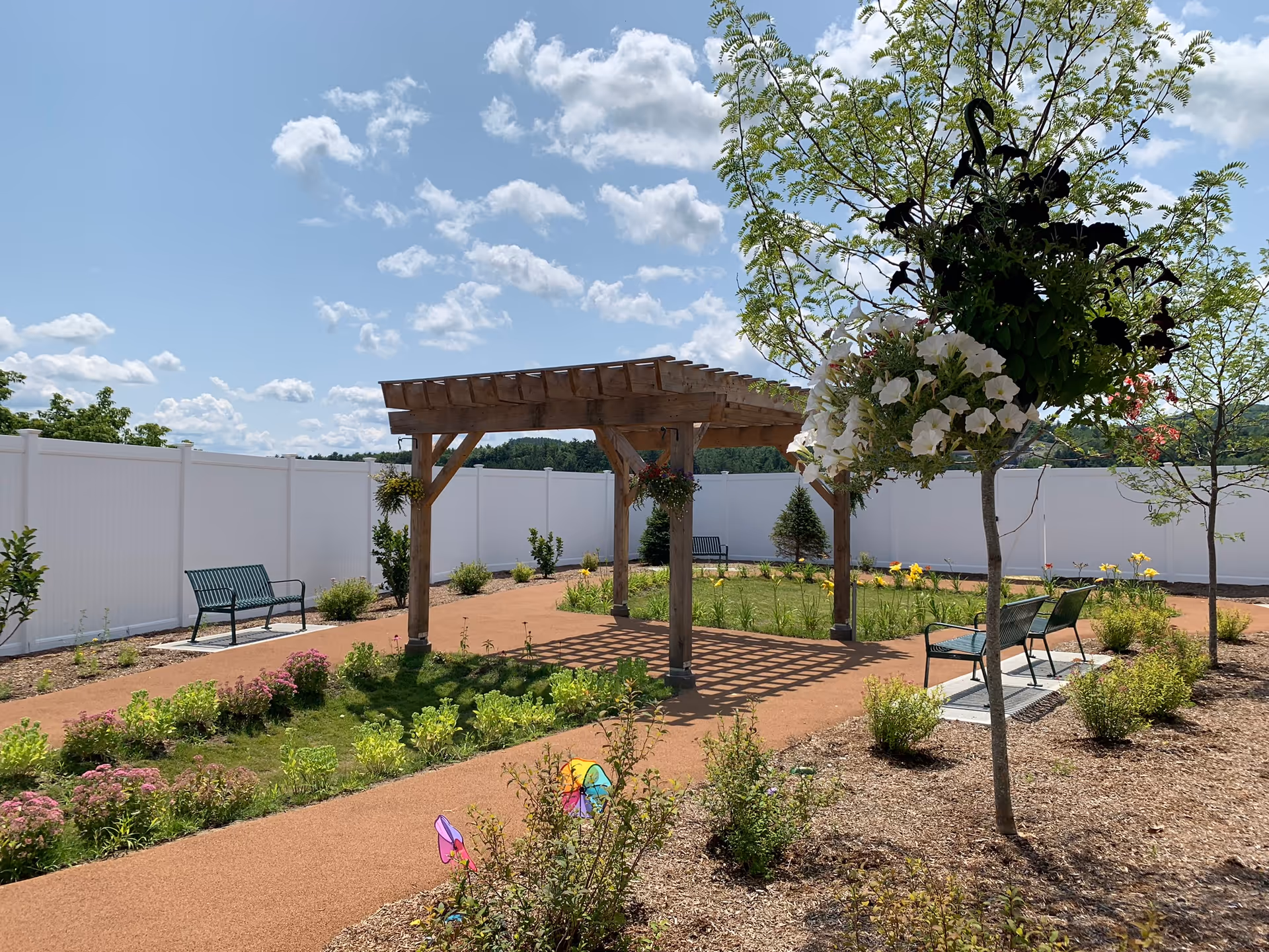 Outdoor garden area with a wooden pergola, green benches, flowering plants, and a white privacy fence under a partly cloudy blue sky.