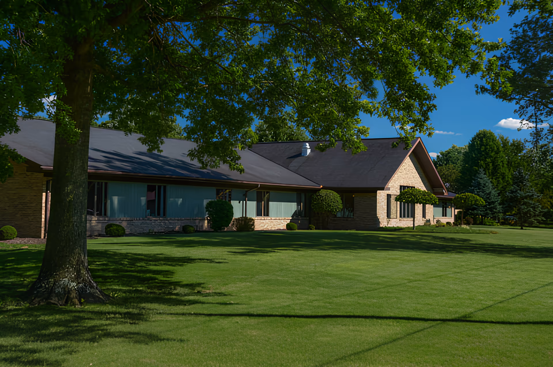 Single-story brick health care building with a wide green lawn and large trees in front under a blue sky.
