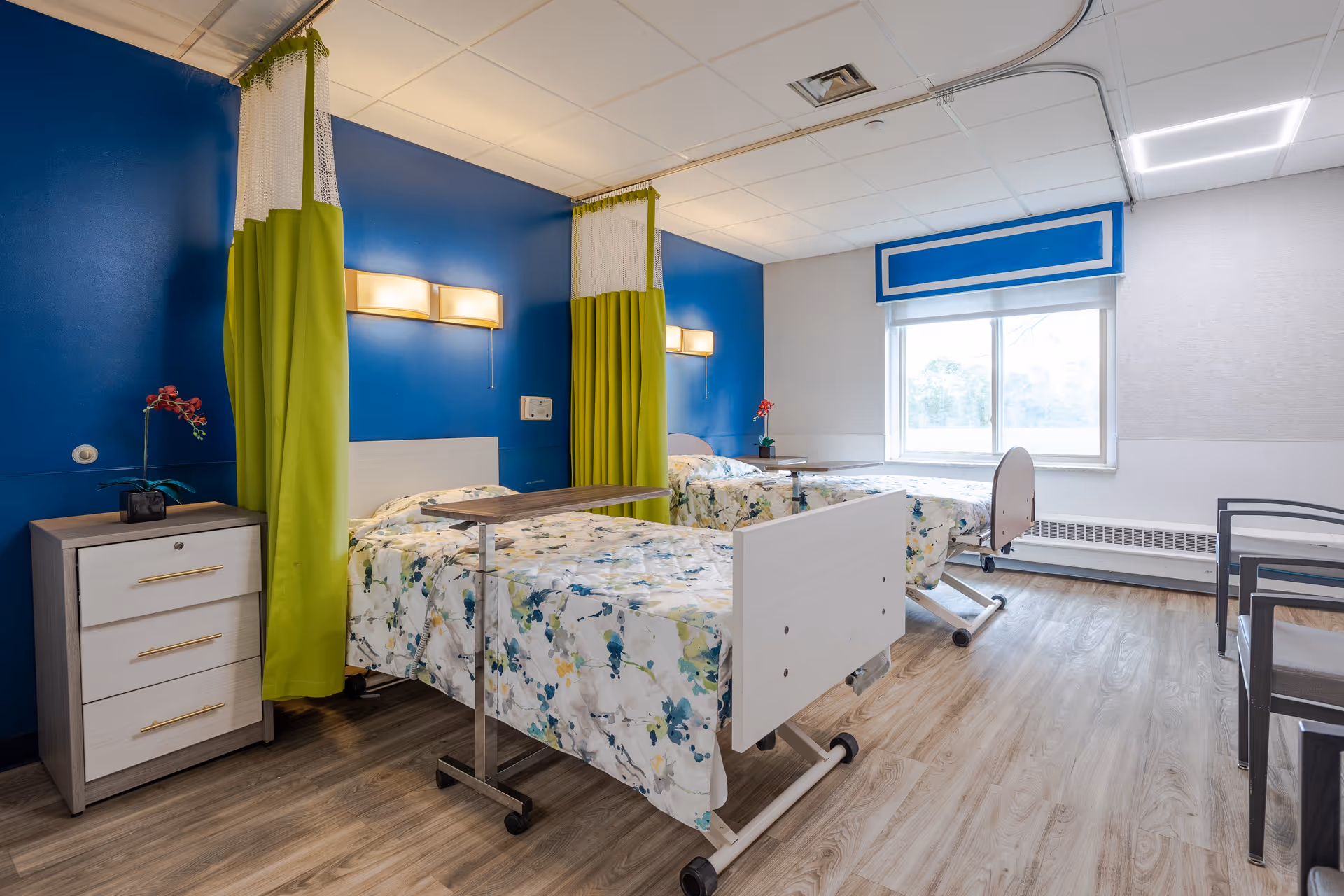 Two beds with blue walls, green privacy curtains, and bedside furniture in a shared patient room.