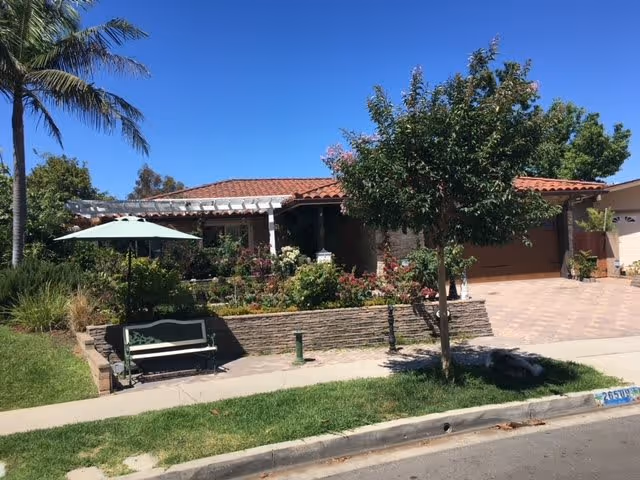 Front exterior view of a single-story house with a red tile roof, a garage, a garden with various plants and flowers, a tree, a bench with an umbrella, and a palm tree under a clear blue sky.