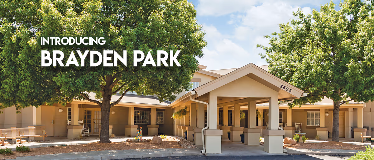 Exterior view of Brayden Park Assisted Living & Memory Care facility showing a beige building with a covered entrance, surrounded by trees and landscaping under a partly cloudy sky.