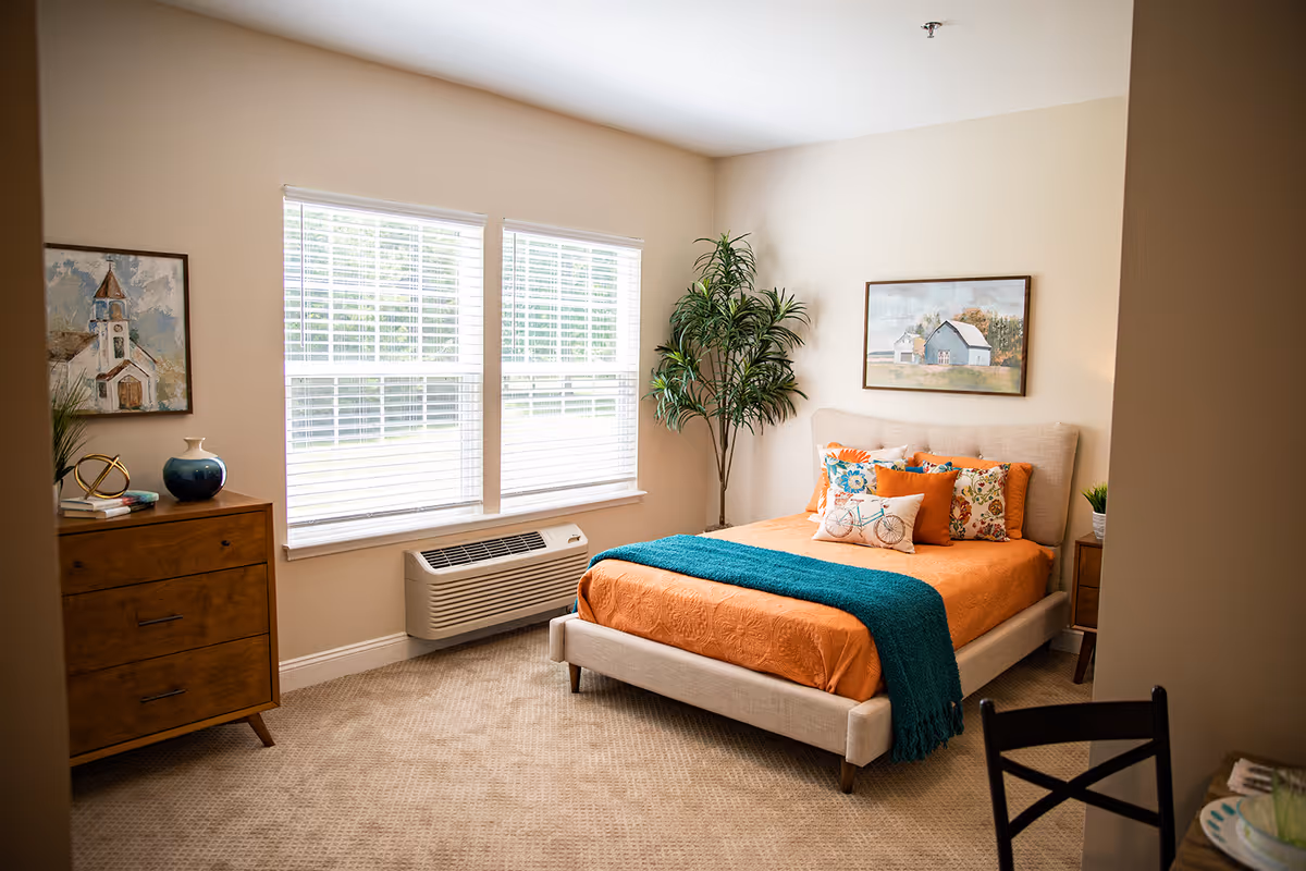 A cozy bedroom with a beige upholstered bed dressed in an orange bedspread and colorful pillows, a teal throw blanket, a wooden dresser with decorative items, a potted plant in the corner, two large windows with white blinds, and framed artwork on the walls.